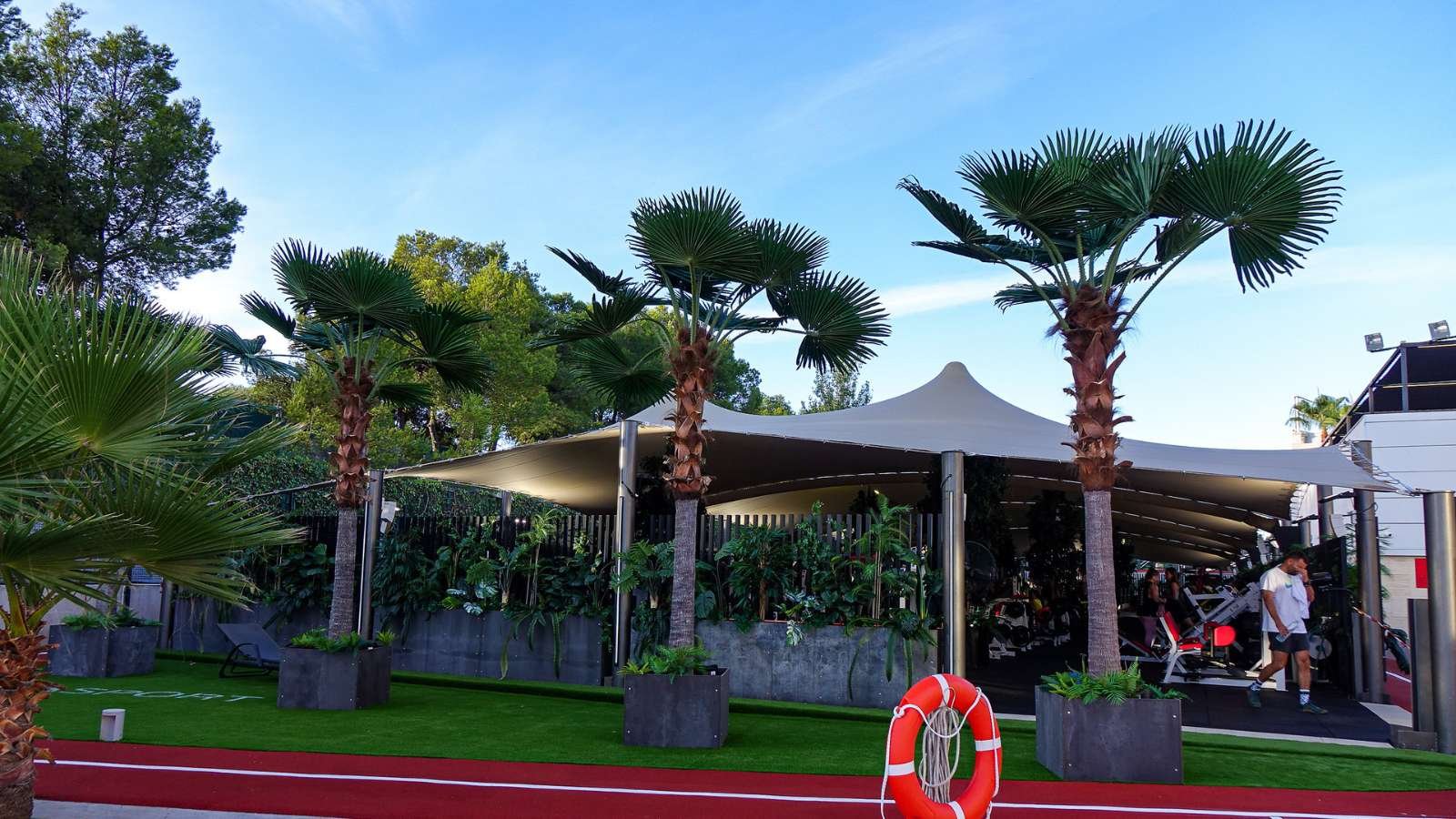 A row of highly realistic artificial fan palm trees separating a red running track from a poolside sun lounger area under a bright blue sky.