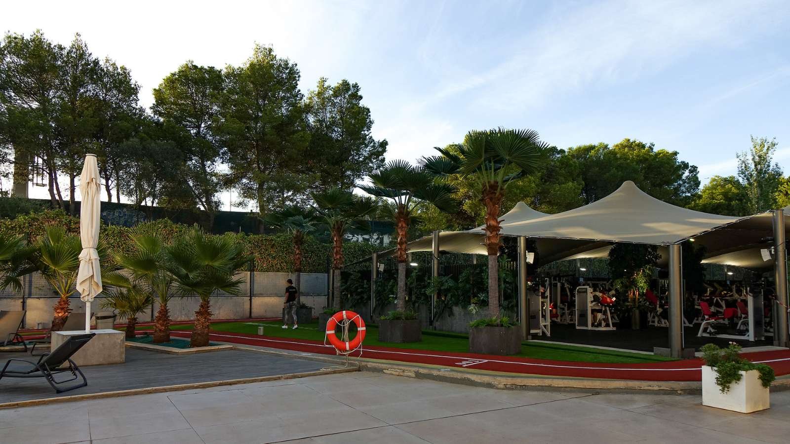 A different angle of the sports complex, showing the line of artificial palm trees running parallel to the track and pool area, emphasizing the contrast with the red and green turf.